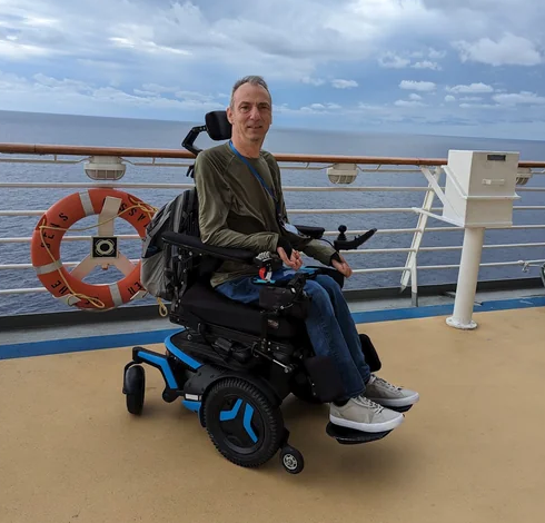 Kelly Michels on the open deck of a cruise ship in a power wheelchair, smiling, sea and cloud behind him